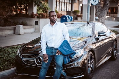 Fortune Vieyra dressed in business casual clothing and sitting on the hood of a Mercedes with hand on jeans in the city..